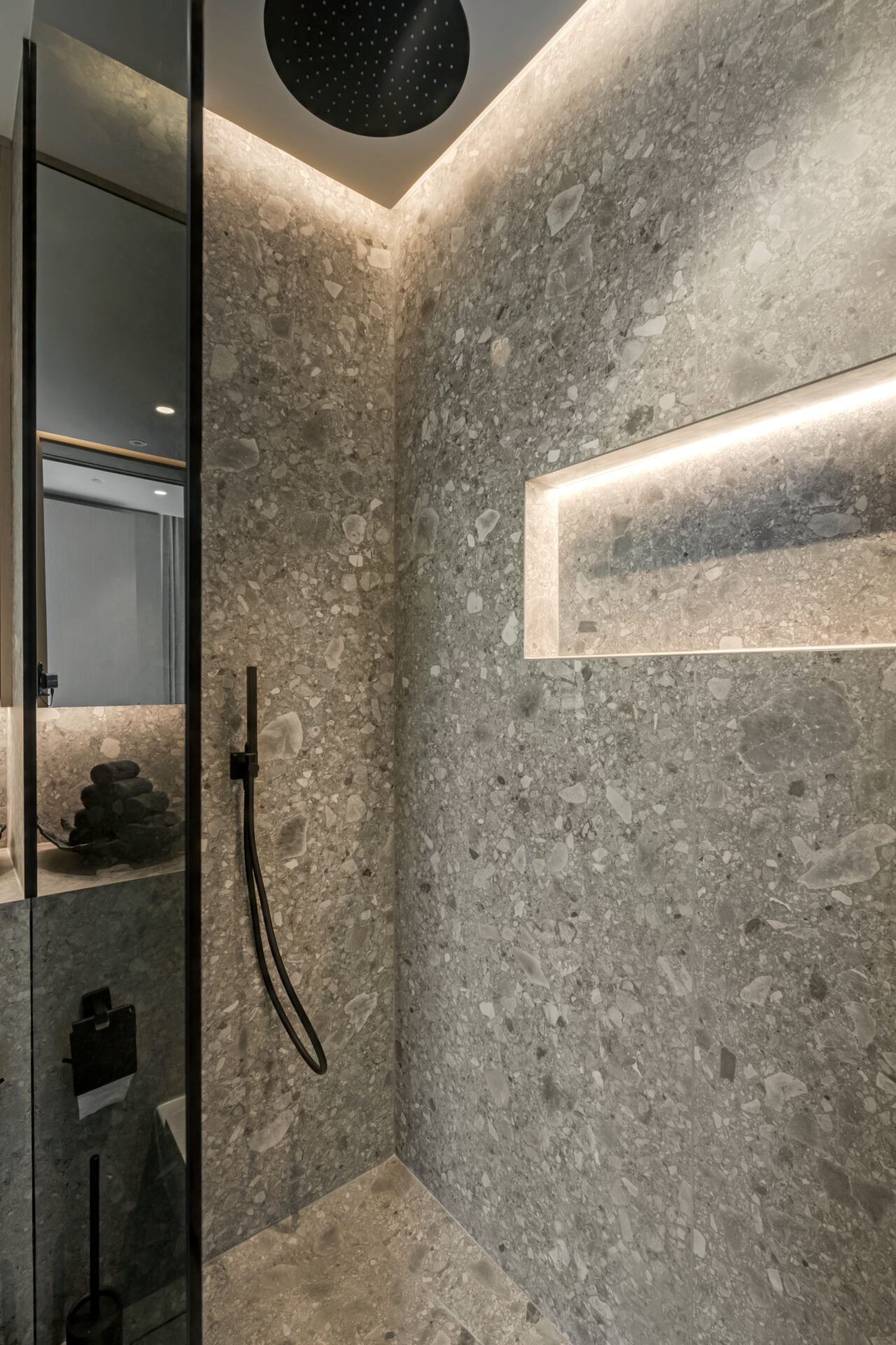 Luxury walk-in shower with grey terrazzo stone walls, recessed LED-lit niche, black rainfall showerhead, and handheld shower.