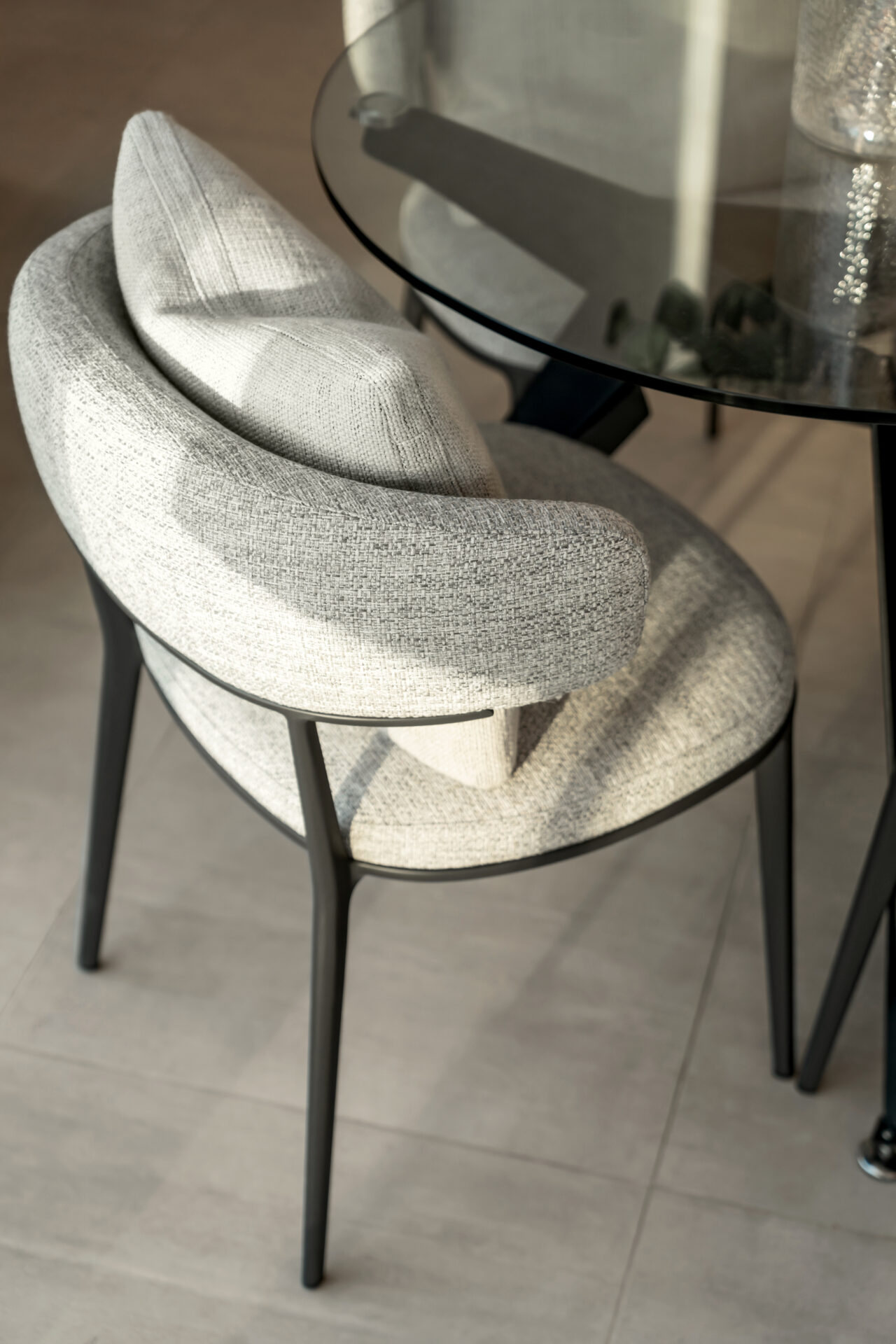 Close-up of a modern upholstered dining chair in light grey fabric with a curved backrest and black metal legs, next to a glass dining table.