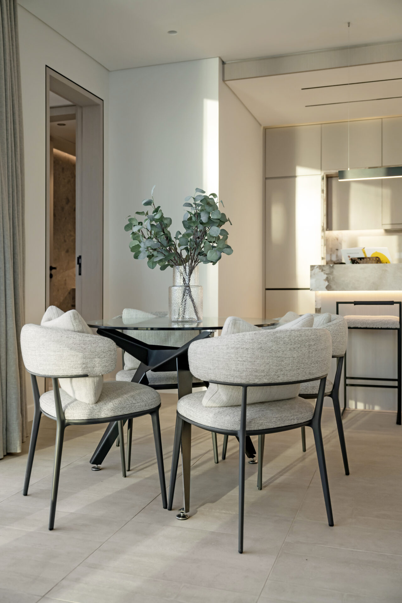 Modern dining area with upholstered grey chairs, glass table, and decorative greenery in a glass vase.